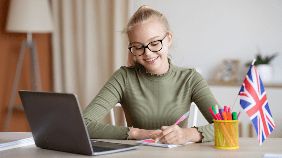 Bir masa başında, dizüstü bilgisayarının karşısında oturan, gözlüklü ve sarı saçlı genç bir kadın defterine not alıyor. Masada renkli kalemler ve bir İngiltere bayrağı bulunuyor.